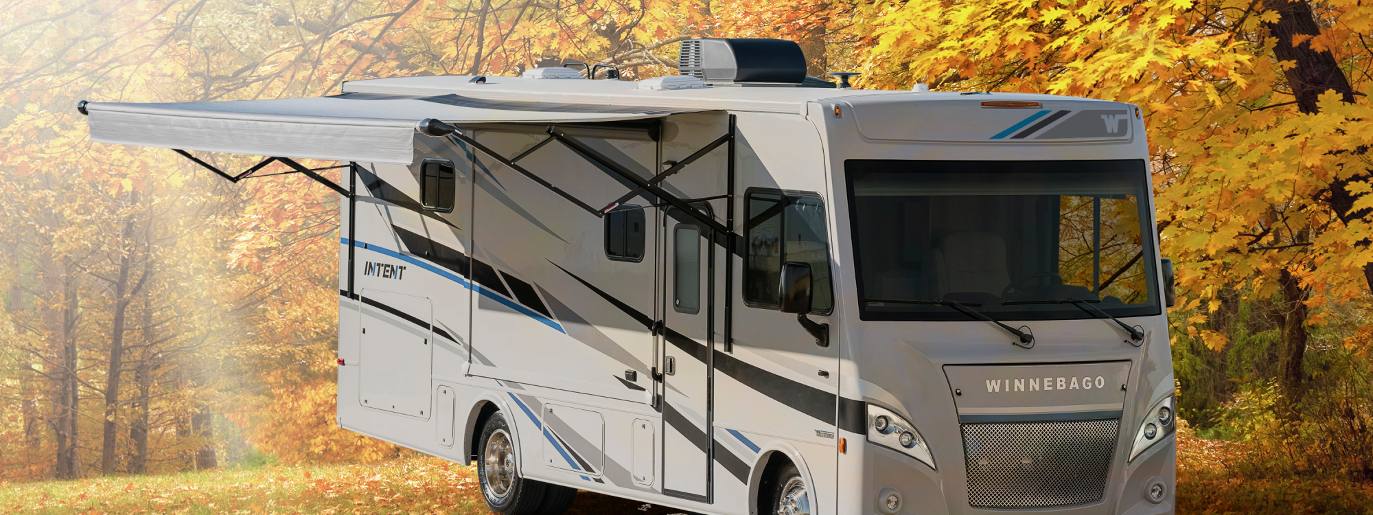 Winnebago Intent exterior with awning fully extended, parked in a wooded area during autumn
